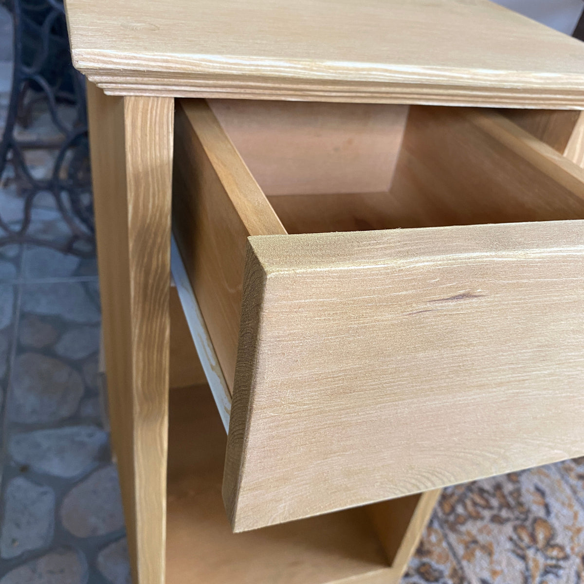 A light natural wood side table or cabinet with an open drawer and open shelf, photographed in a real indoor setting with tiled floor and patterned rug visible.
