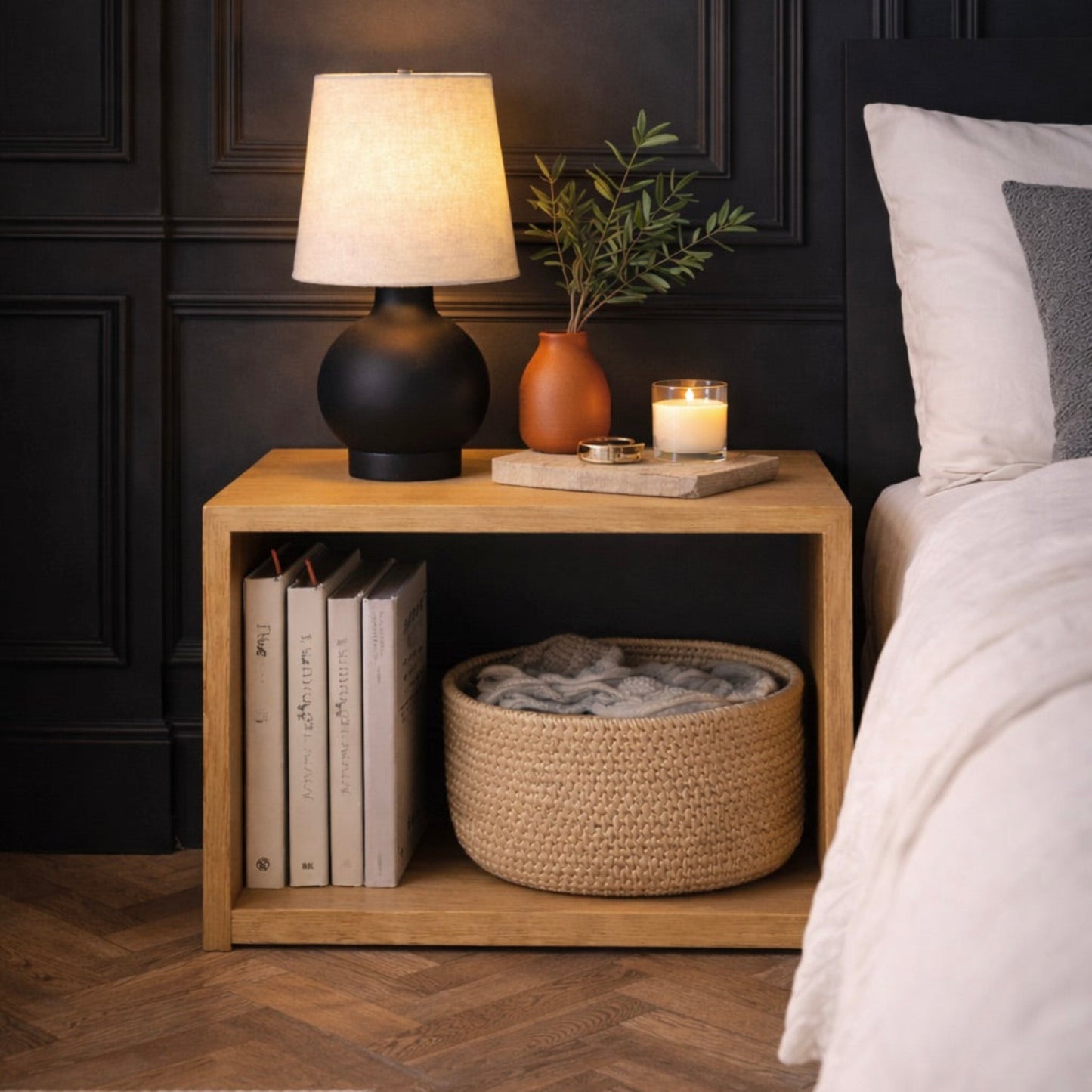 Wooden nightstand with lamp, books, and candle next to a bed in a bedroom.