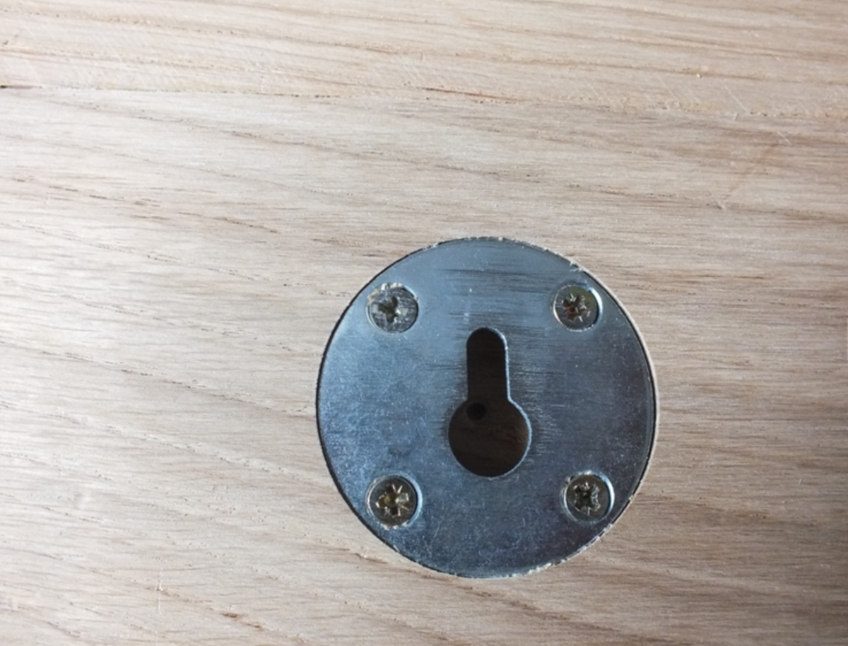 A round metal keyhole cover plate with four mounting screws, shown on a light wood grain surface.