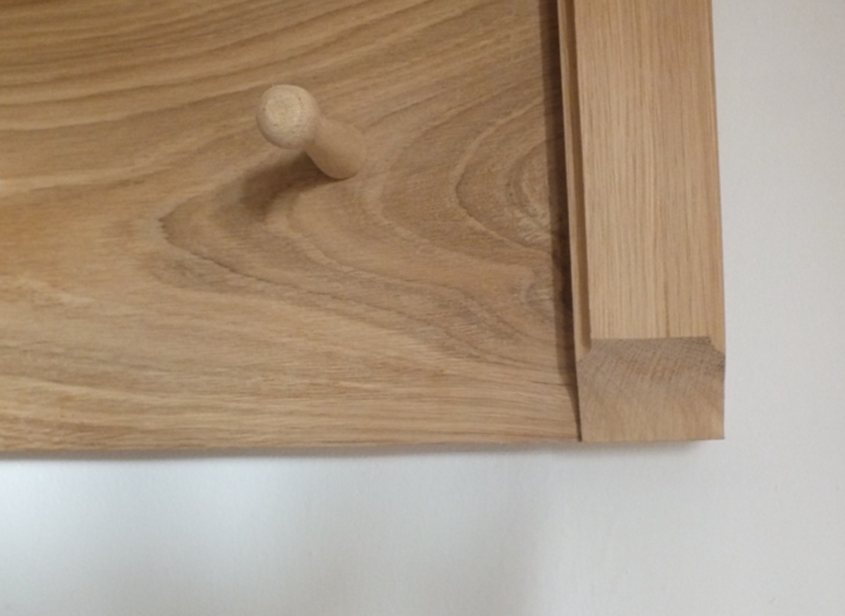 Close-up of a light natural wood wall-mounted coat rack with round peg hooks and a vertical side frame, shown against a plain white wall.