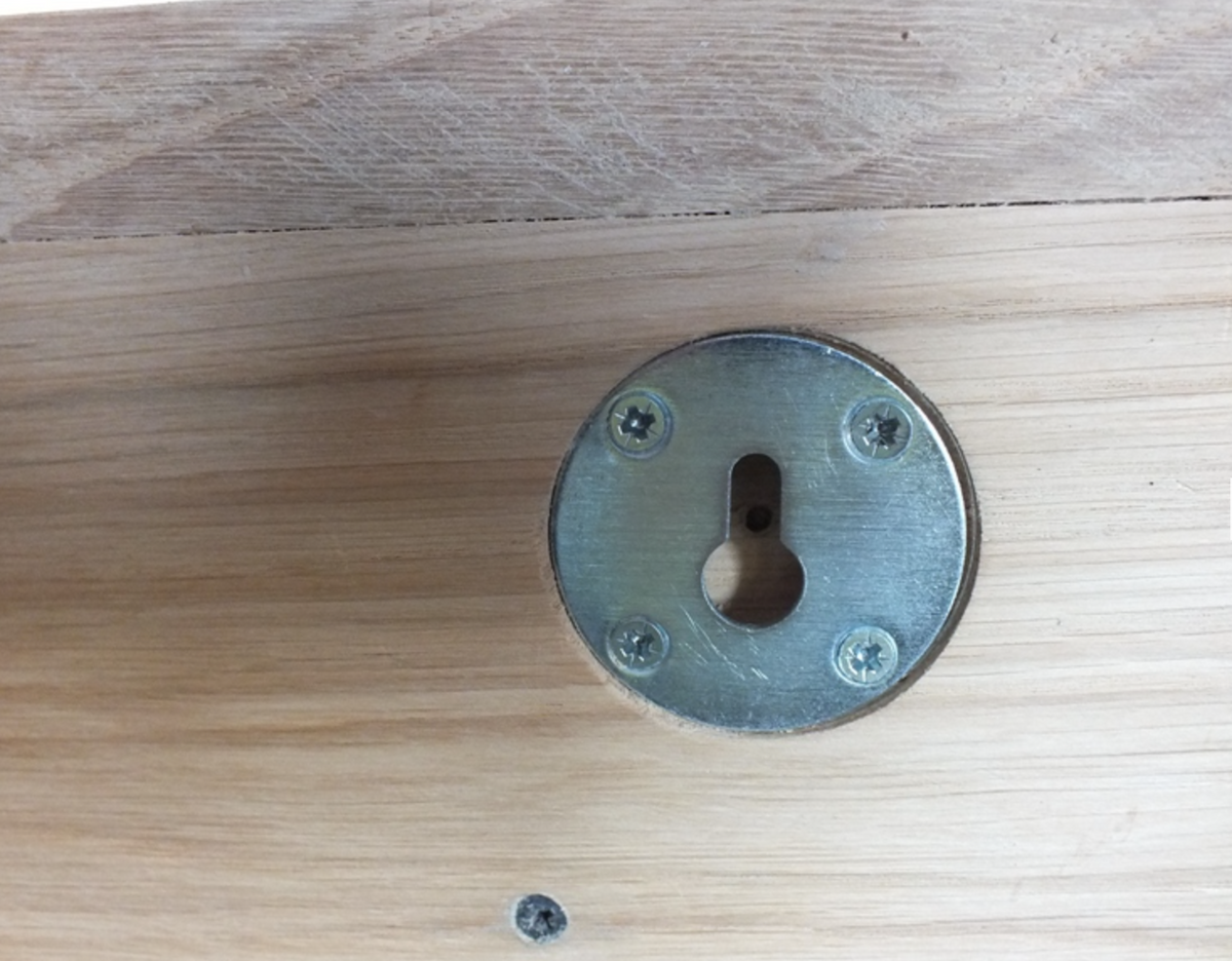 A round metal keyhole cover plate with four screws, mounted on a light-colored wooden surface.