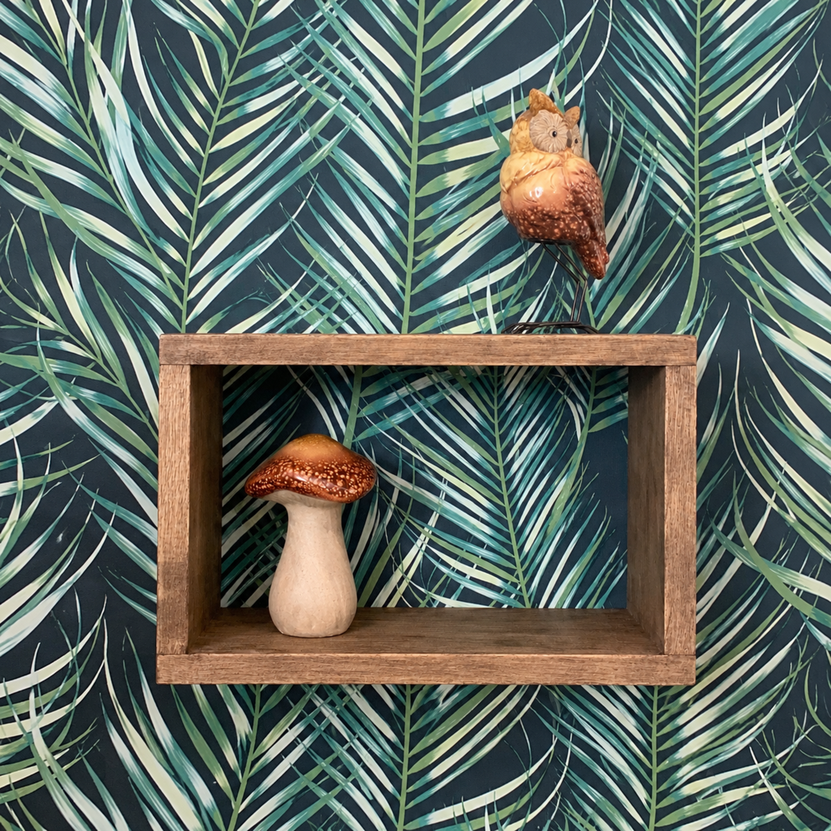 A rustic wooden floating shelf mounted on a dark green and teal palm leaf patterned wallpaper. The shelf contains a small ceramic mushroom figurine, and a small bird figurine sits on the top edge of the shelf.