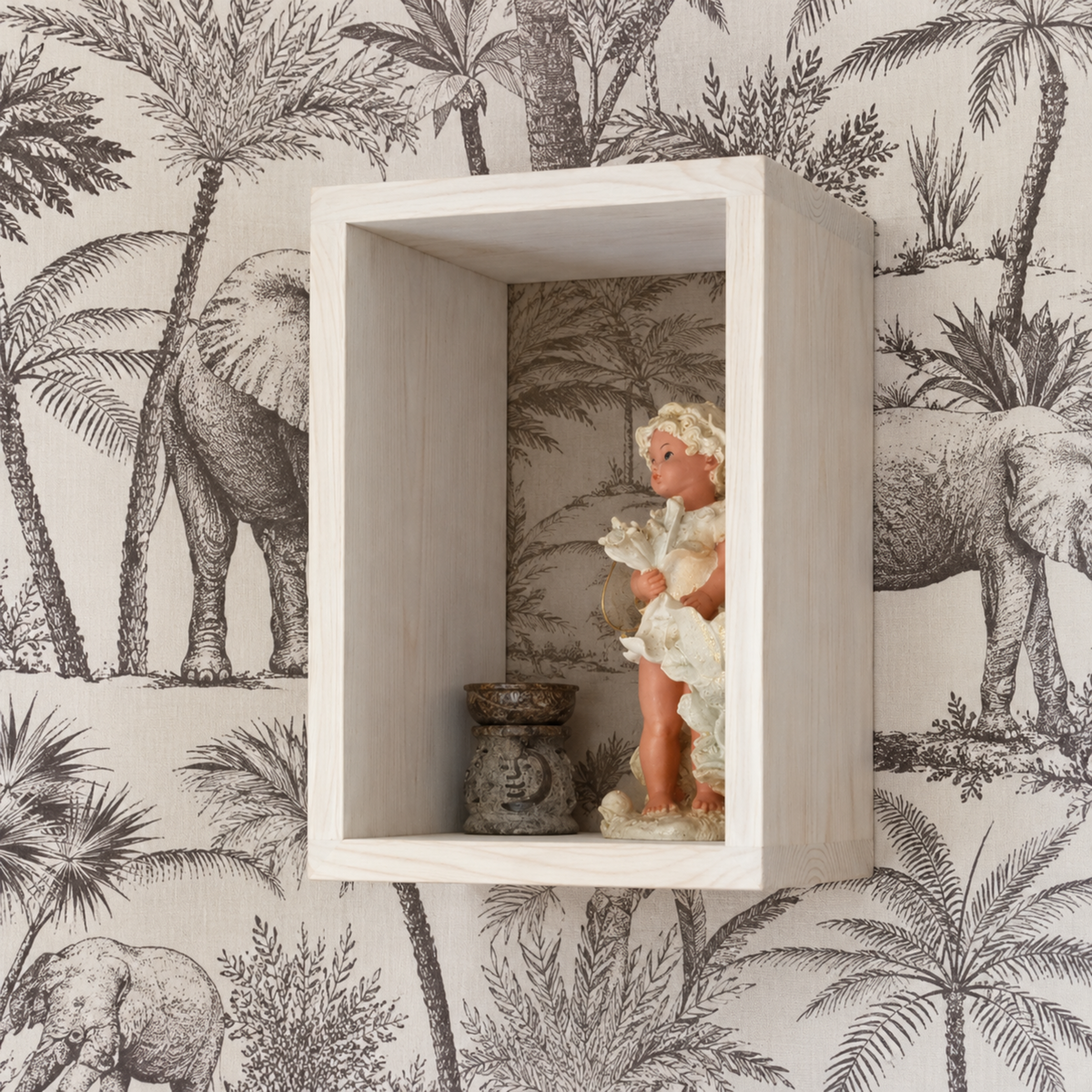 A rectangular white wooden floating shelf mounted on a black and white elephant and palm tree patterned wallpaper. The shelf contains a small decorative figurine of a child in white clothing and a small dark metal bowl.