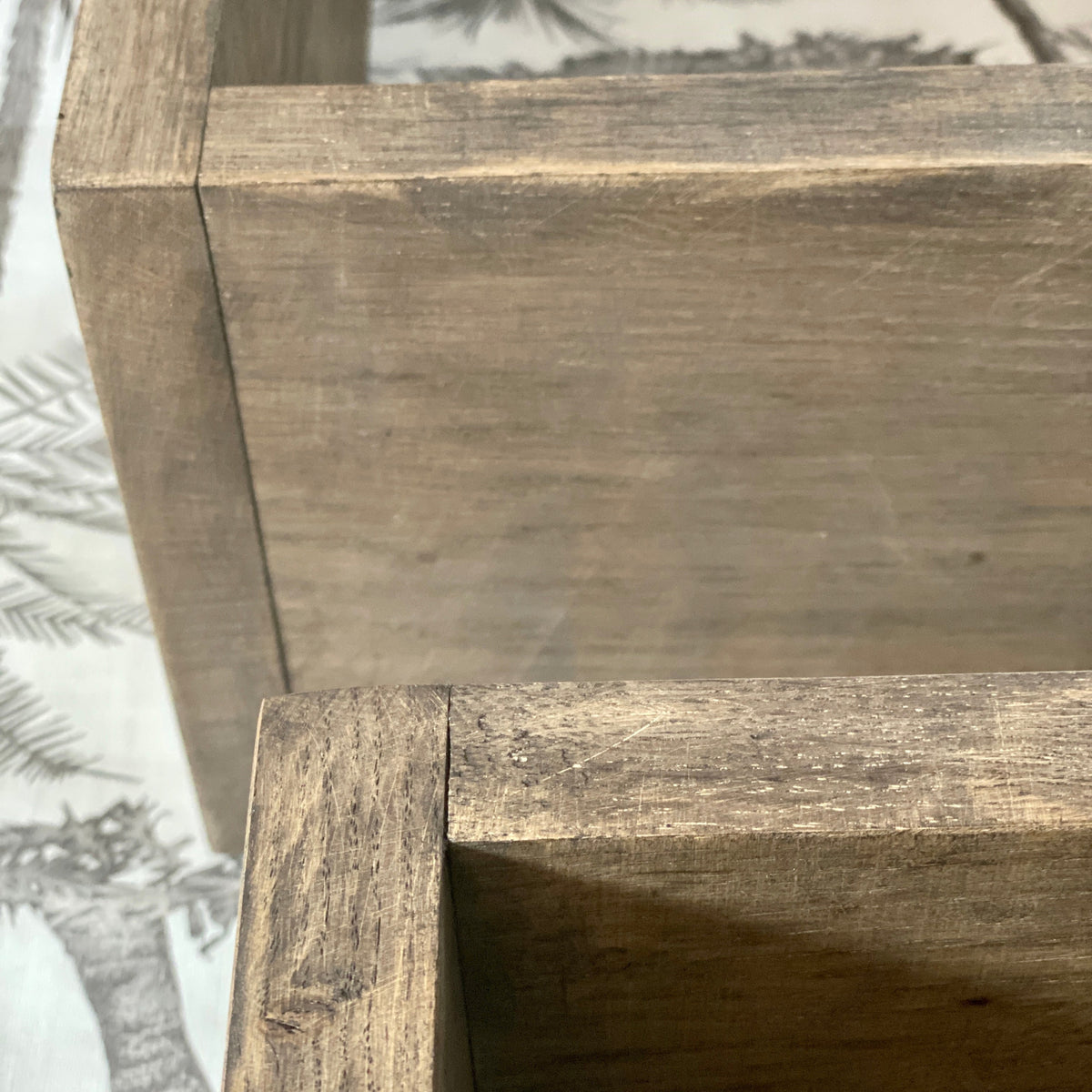 Close-up of a rustic wooden shelf or box corner with weathered brown wood grain and visible seams, photographed against a patterned surface.