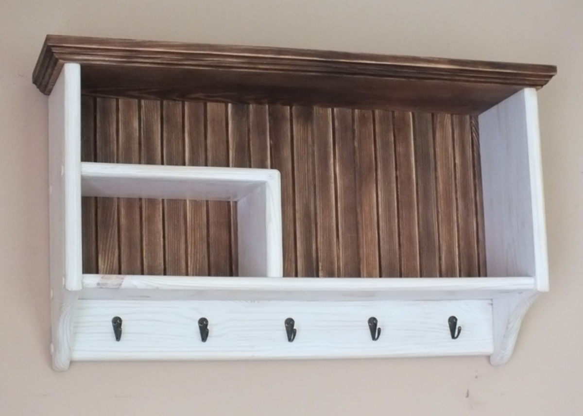 A rustic wall-mounted wooden shelf with a dark brown stained top and slatted back panel, white side and lower frame, a built-in open cubby, and five black hooks along the bottom, shown against a plain beige wall.
