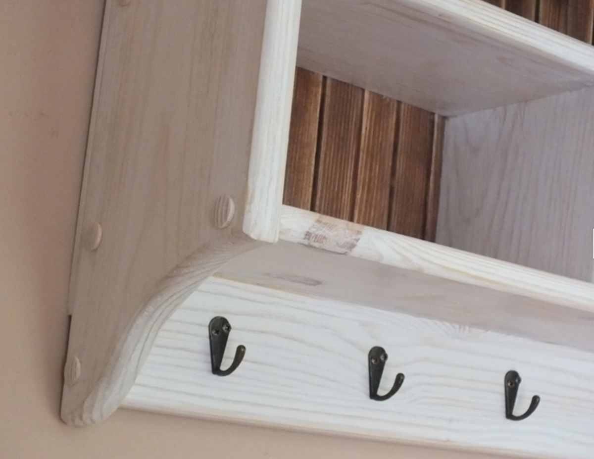A close-up of a light natural wood wall-mounted shelf or cabinet corner with three black metal hooks on the front rail, set against a beige wall.