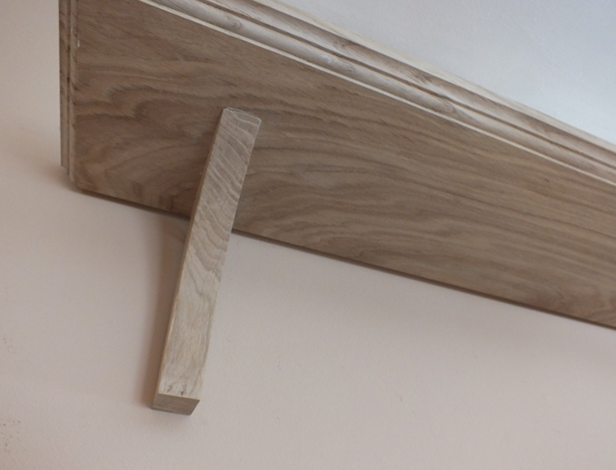 A close-up of a wooden shelf bracket with a light oak wood top and a pale wood support leg, mounted against a plain beige wall.