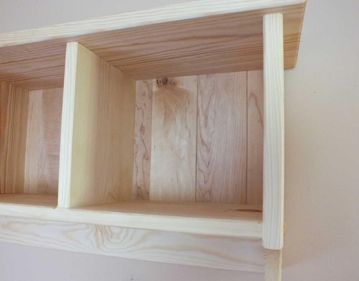 A close-up of a light natural wood wall-mounted shelf with three open cubby compartments, showing the grain and construction details against a plain wall.