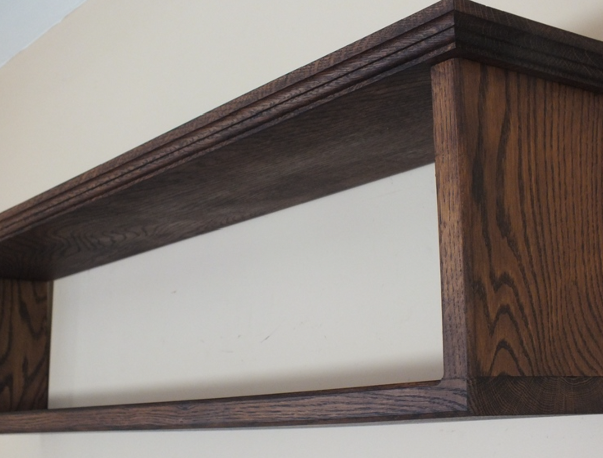 Close-up of a dark brown wooden floating shelf with visible wood grain, mounted against a plain off-white wall.