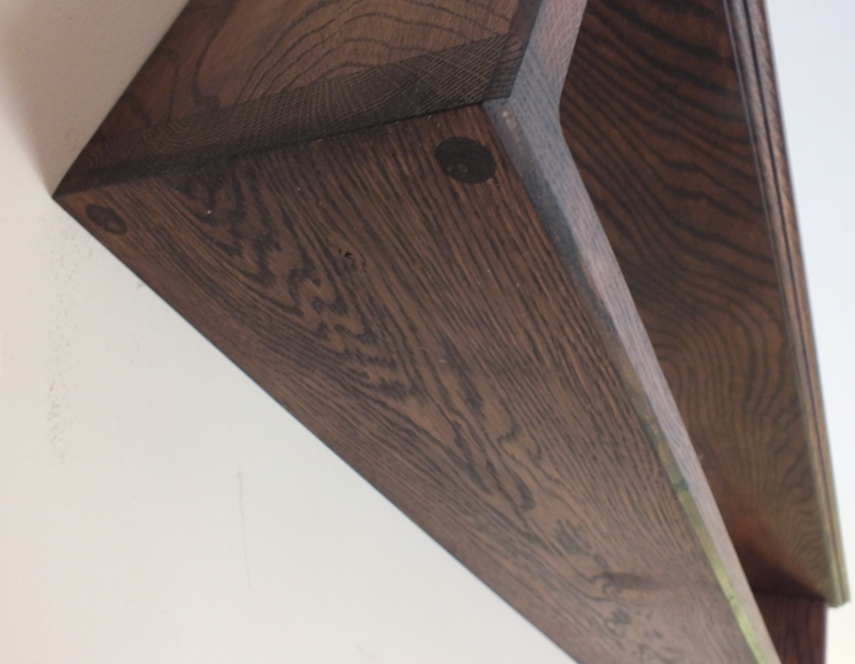 Close-up of a dark brown wooden shelf corner with visible wood grain and a small round mounting hole, photographed against an off-white wall.