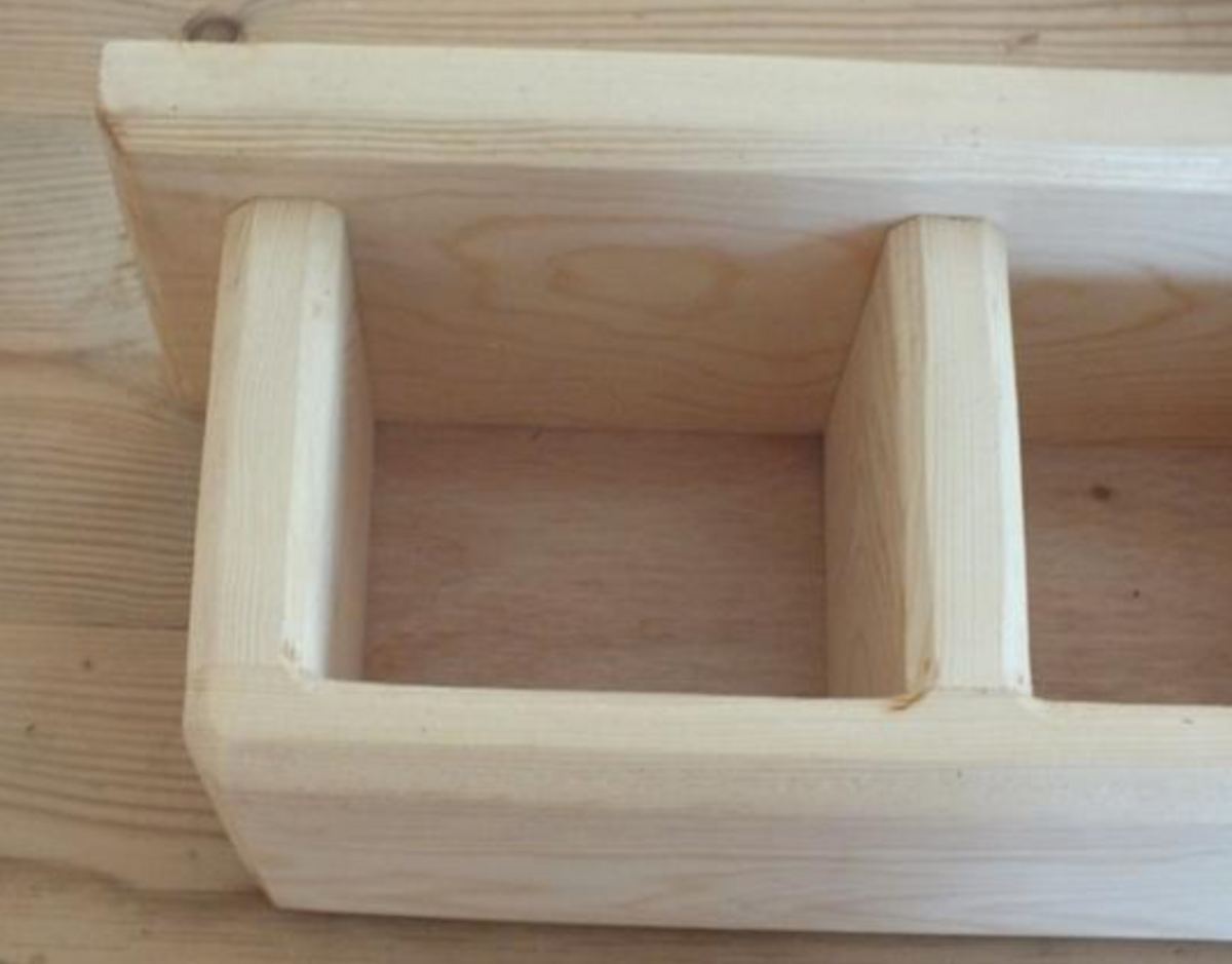A close-up of a light natural wood shelf or organizer with two vertical compartments and a top rail, shown on a matching wood surface.