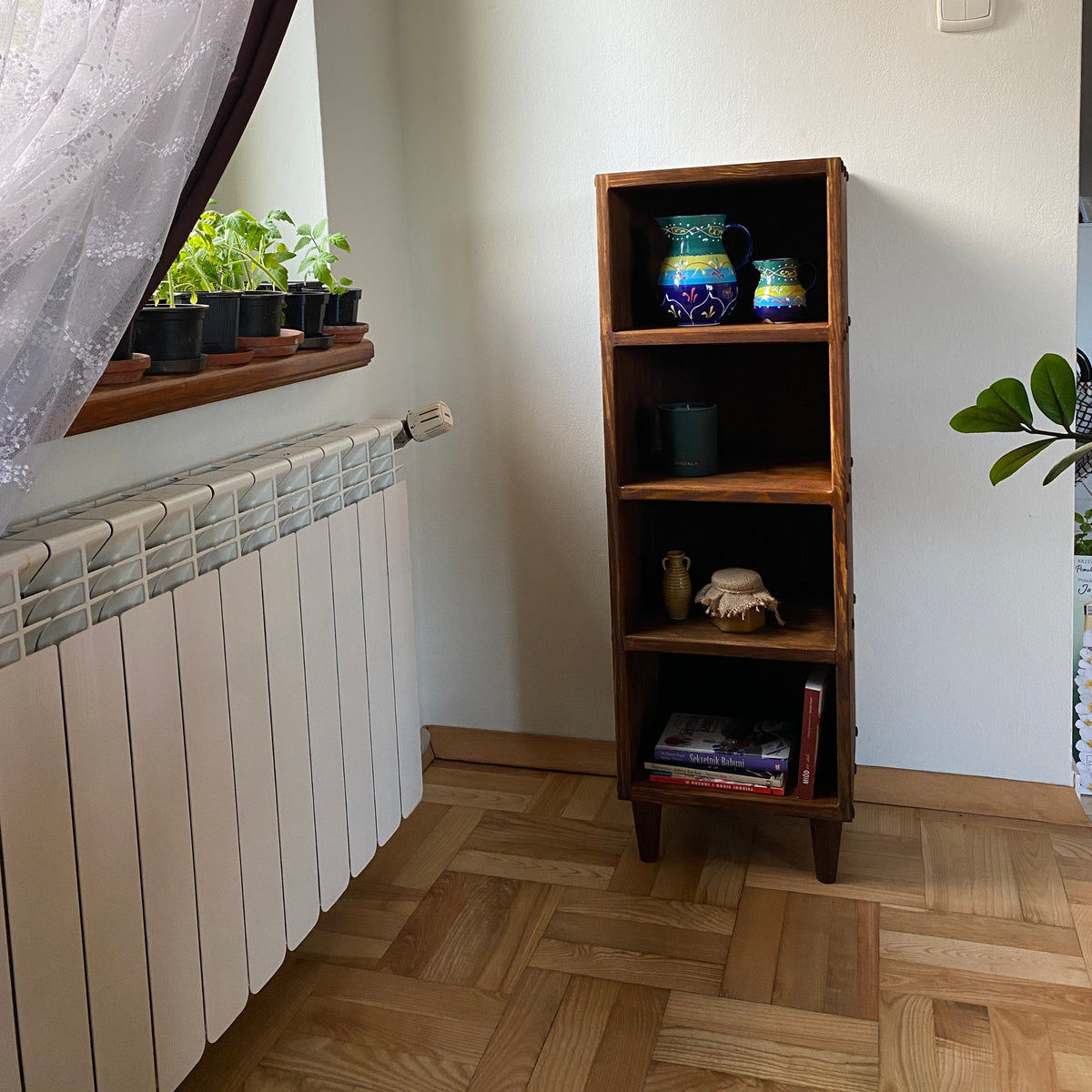 A dark brown wooden four-compartment shelf mounted on a white wall in a room with a parquet wood floor and a white radiator.