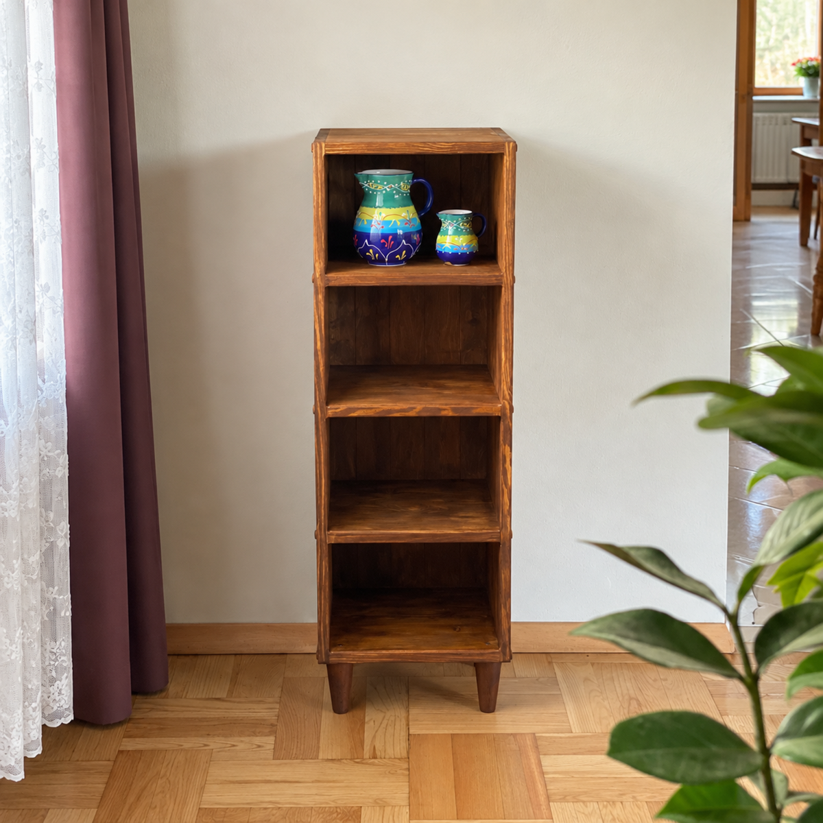 A tall, narrow wooden 4-tier shelf with tapered legs stands on a light wood parquet floor against a beige wall. The shelf is styled with two blue and green ceramic pitchers on the top shelf.