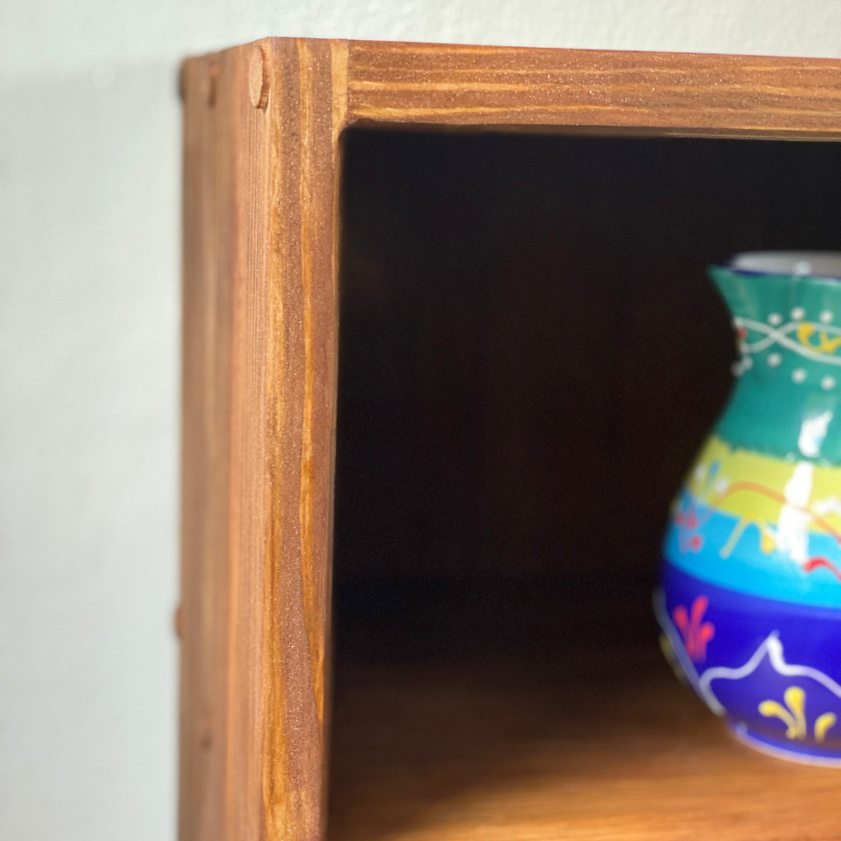 Close-up of a rustic wooden shelf with a colorful hand-painted ceramic vase on the middle shelf against a light wall.