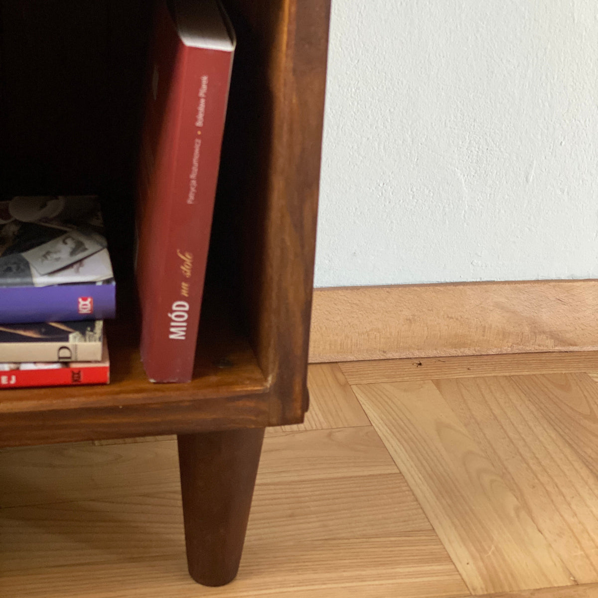 A close-up of a dark brown wooden side table or cabinet leg and shelf, with a red book standing upright inside the open cubby and other books stacked on the lower shelf, set against a white wall and light wood parquet floor.