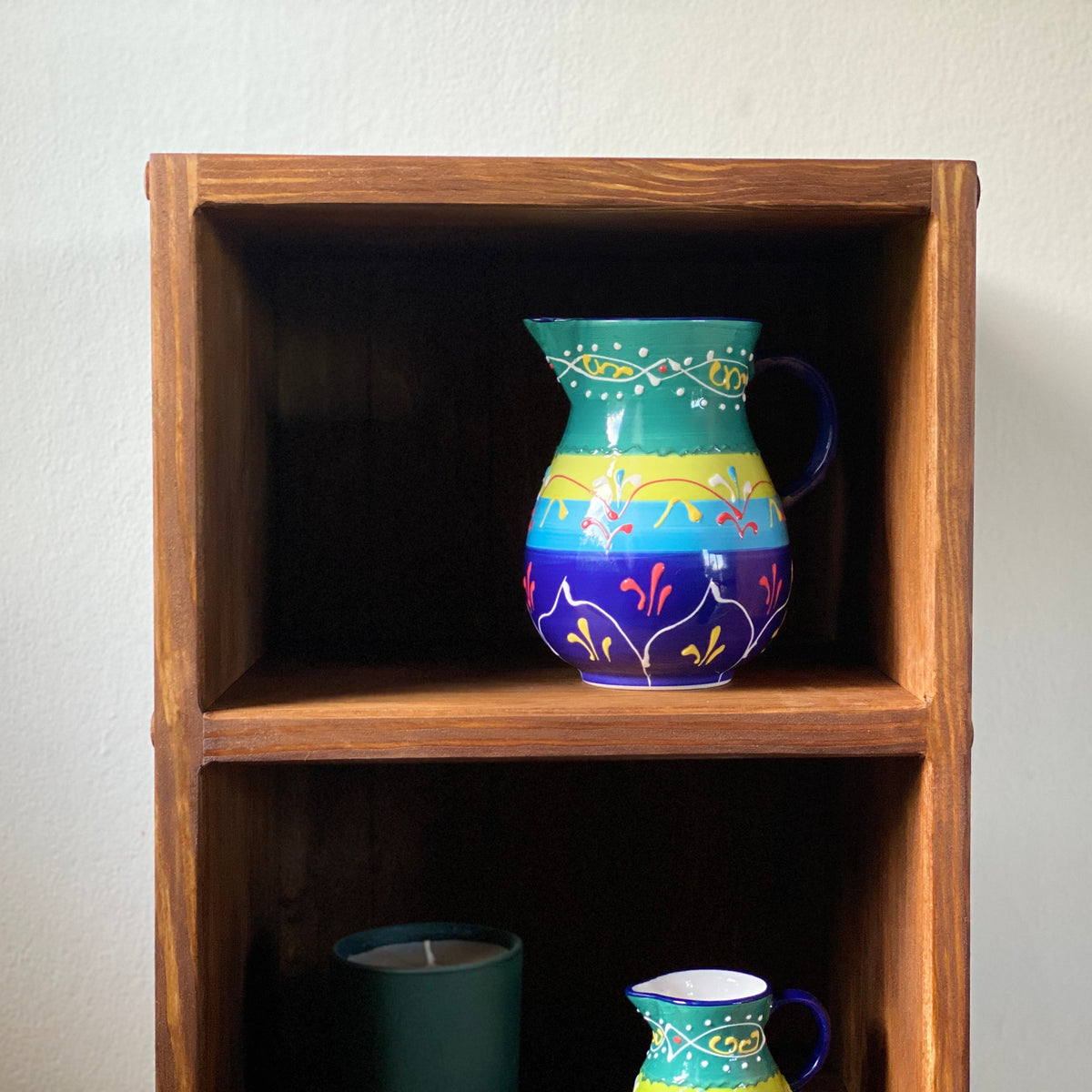 A colorful hand-painted ceramic pitcher displayed on a rustic wooden two-tier shelf against a plain off-white wall.