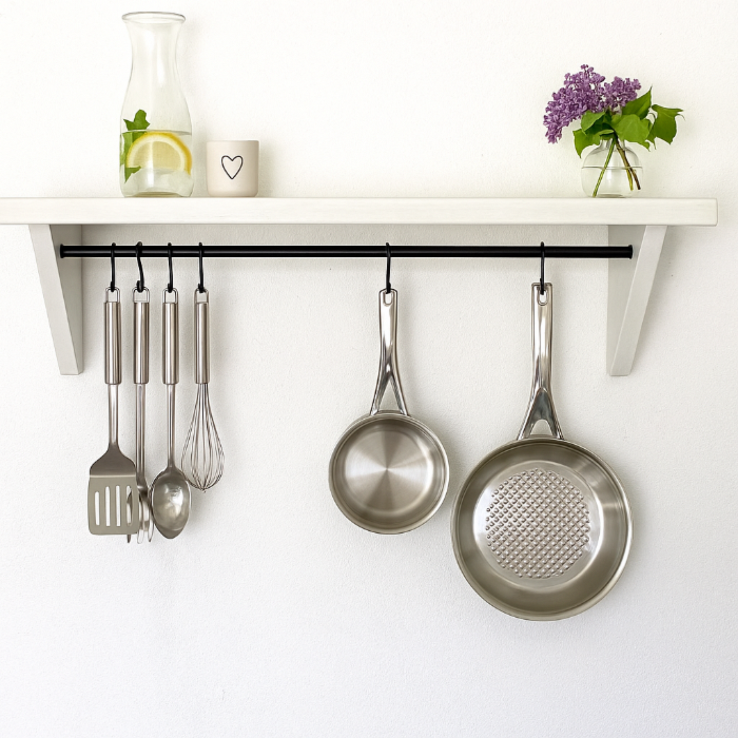 Kitchen utensils and pots hanging on a wall rack with a shelf above containing a carafe, small container, and flowers.