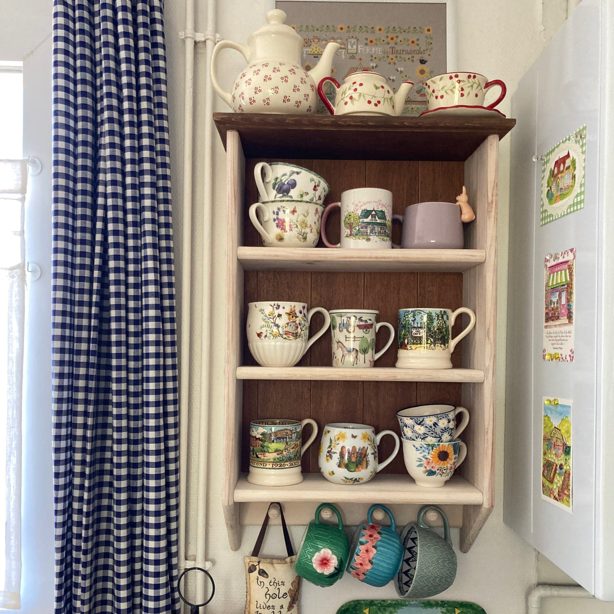 A rustic wooden wall-mounted shelf filled with assorted ceramic mugs, teapots, and cups in a cozy kitchen setting next to a blue-and-white gingham curtain and a white refrigerator.