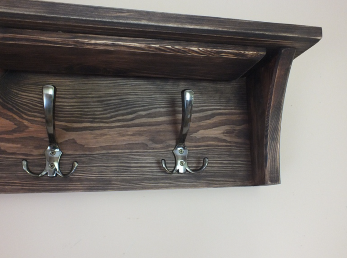 A dark brown wooden wall-mounted shelf with two silver metal coat hooks, shown against a plain off-white wall.