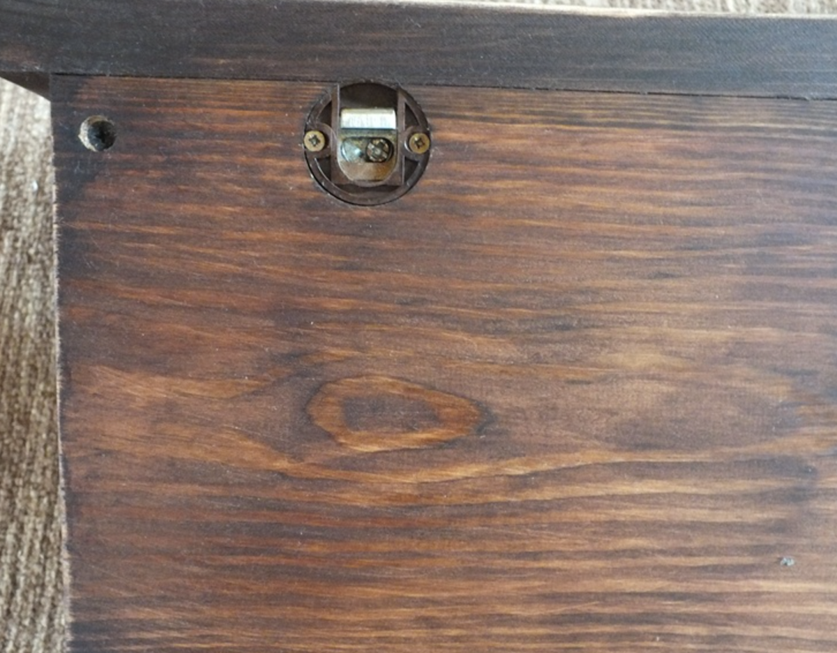 Close-up of a dark brown wooden surface with visible wood grain, a small round metal lock or latch near the top center, and part of a dark wooden edge at the top.