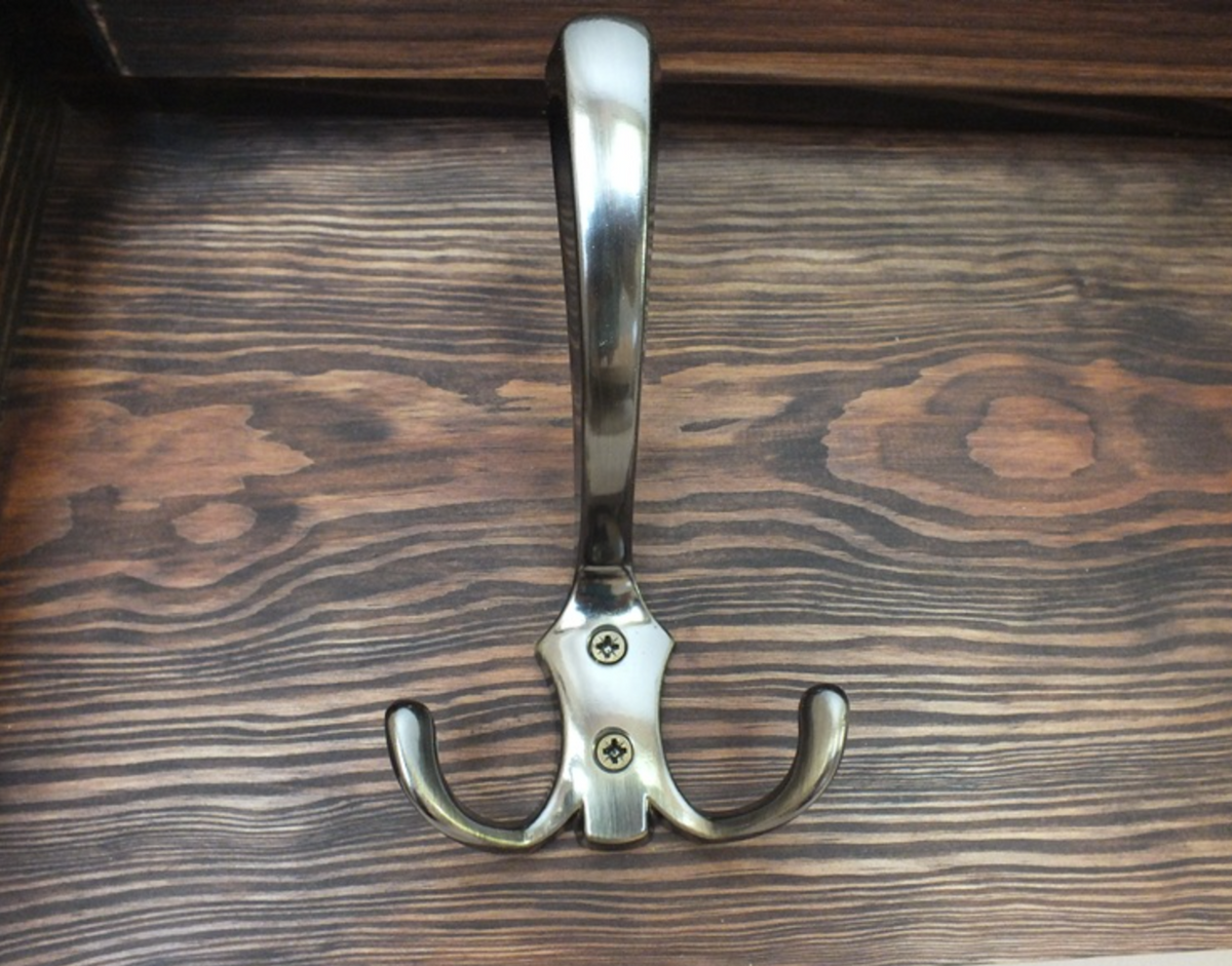 A polished metal double coat rack hook with a single upright hook and one curved hook, mounted on a dark wood surface.