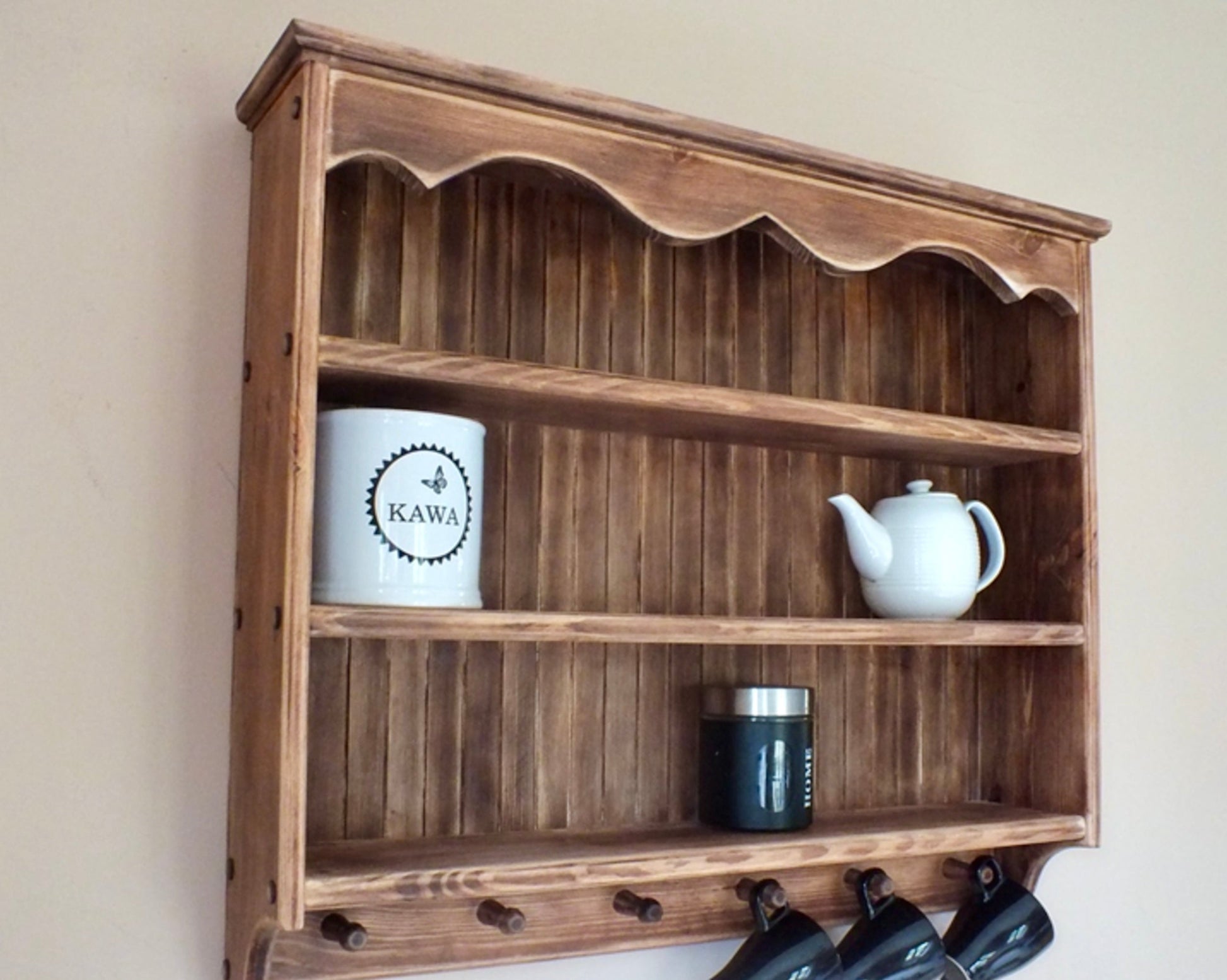 A rustic wooden wall-mounted shelf with three open compartments and hanging hooks at the bottom, shown against a plain beige wall with decorative items placed on the shelves.