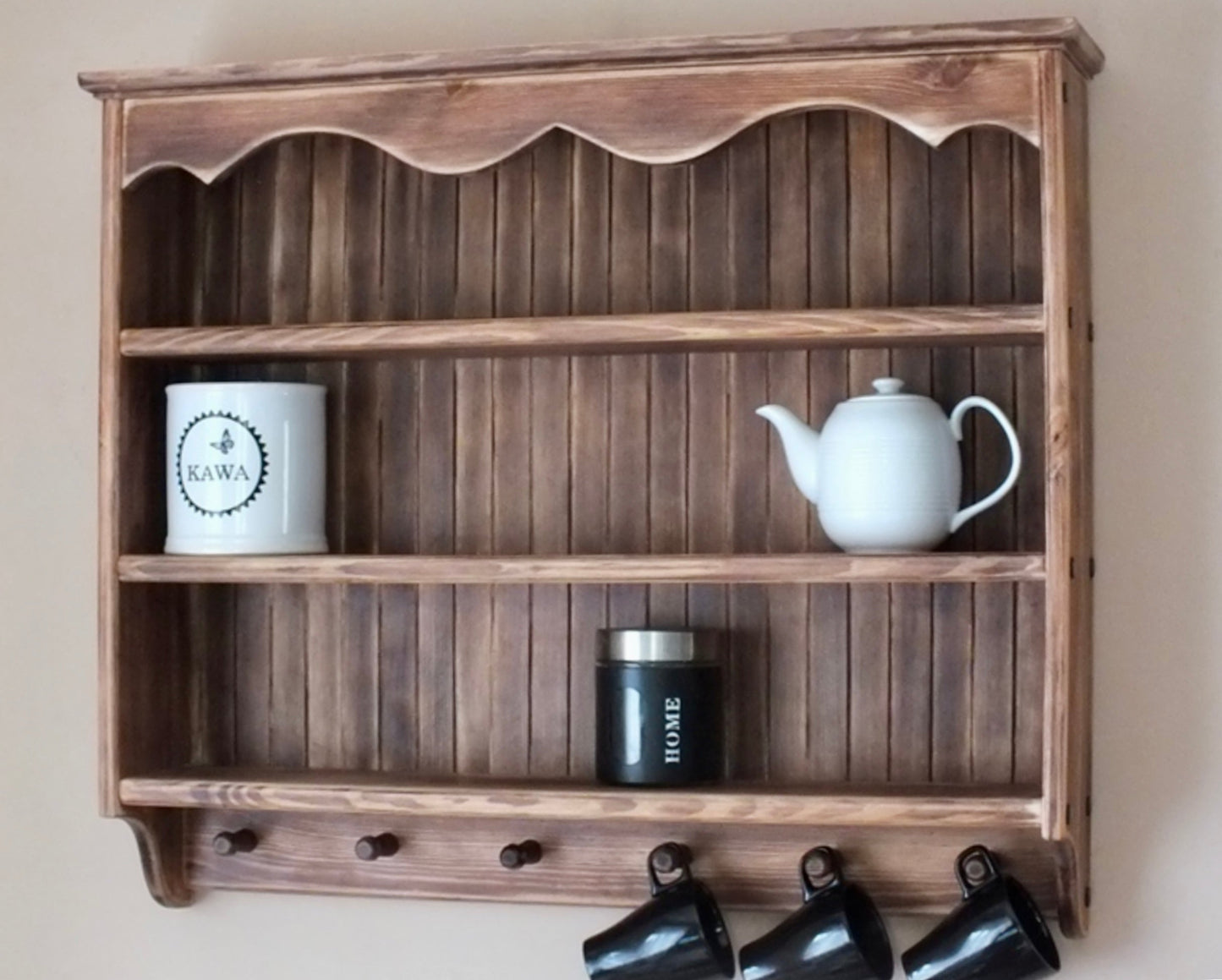 A rustic wooden wall-mounted shelf with three tiers and hanging pegs, styled with a white teapot, a white mug, and a black canister against a beige wall.