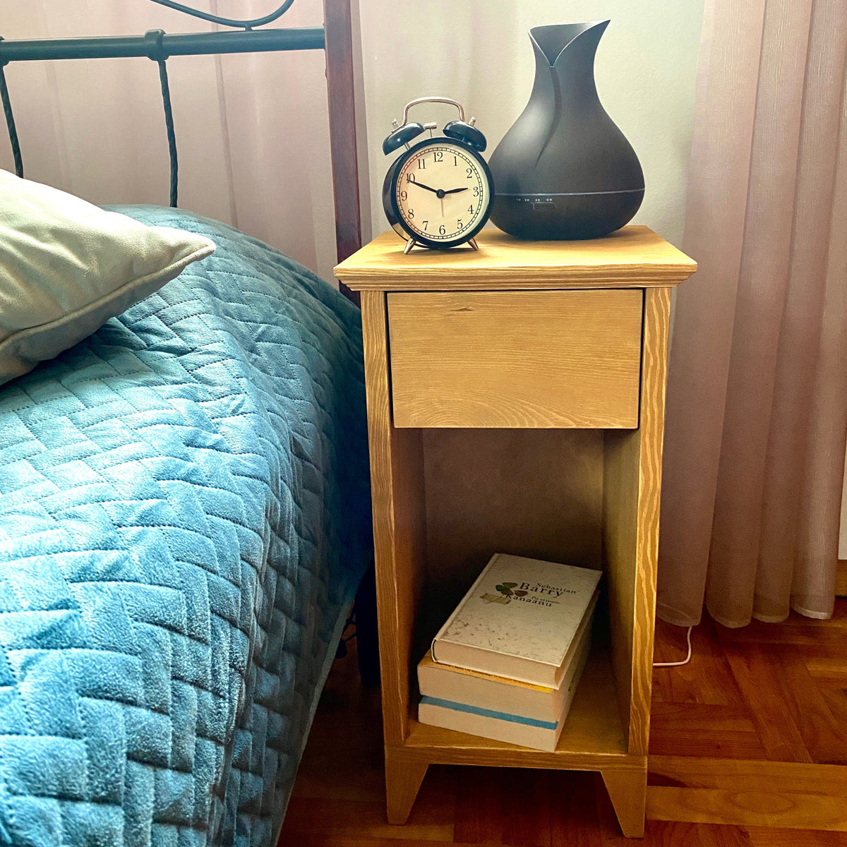 A light natural wood nightstand beside a bed with a blue quilted cover, holding a black alarm clock, a matte black diffuser, and two stacked books on a wood floor.