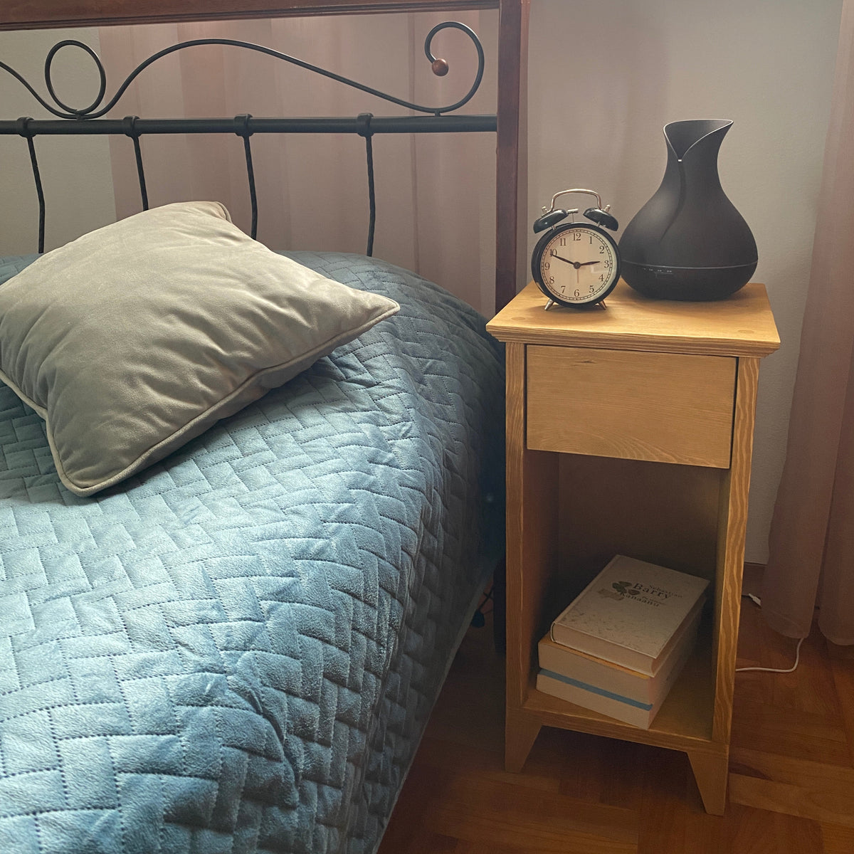 A cozy bedroom corner featuring a light wood nightstand next to a bed with a blue quilted cover and a gray pillow. The nightstand holds a black vase and a vintage-style alarm clock, with books stacked on the lower shelf.