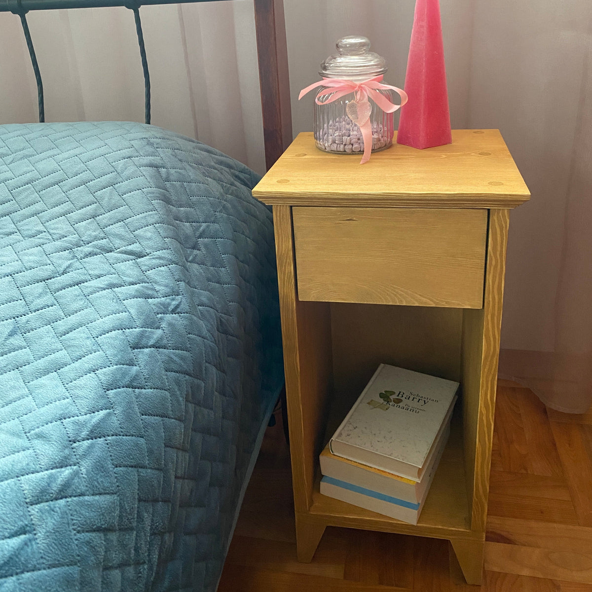 A light natural wood nightstand with one drawer and an open shelf, placed next to a bed with a blue quilted cover. On top of the nightstand are a pink taper candle and a glass jar with a pink ribbon.