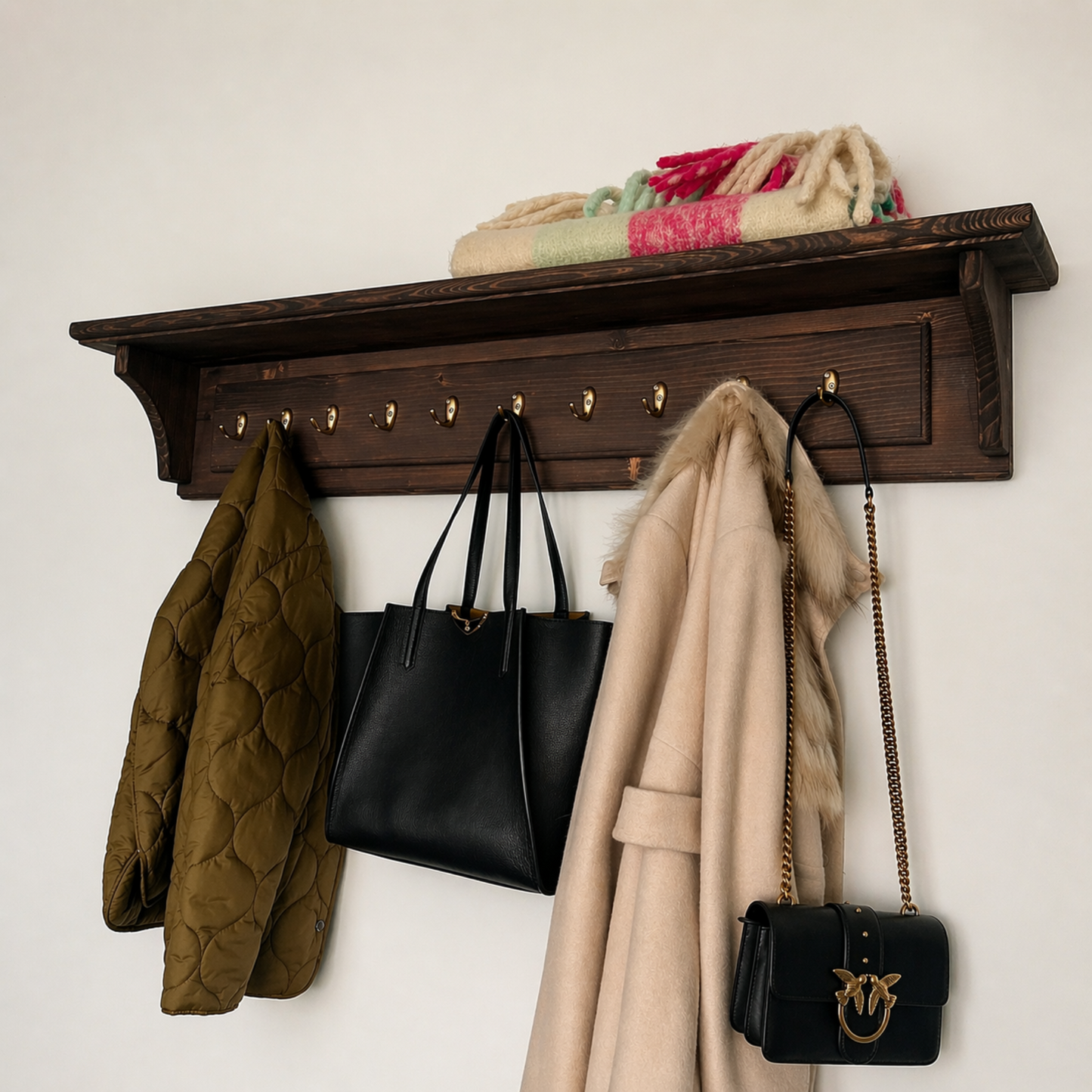 Wooden coat rack with hooks holding a green coat, black handbag, beige coat, and black handbag against a white wall.