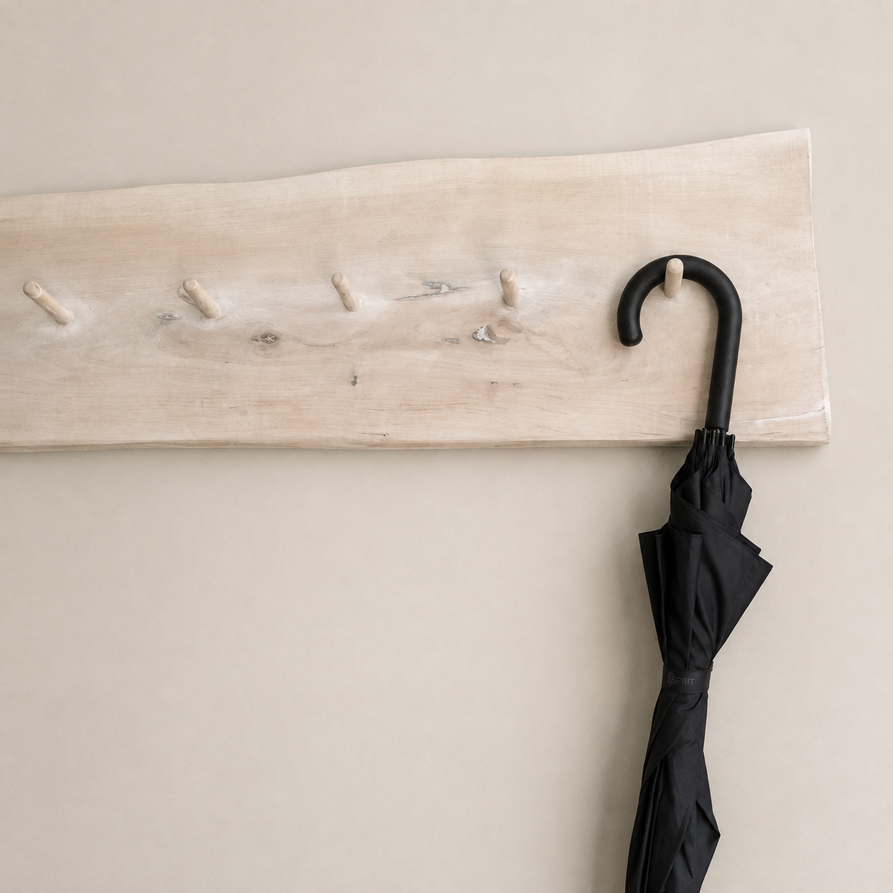A light natural wood wall-mounted coat rack with five pegs and a black umbrella hanging from one hook, shown against a plain beige wall.
