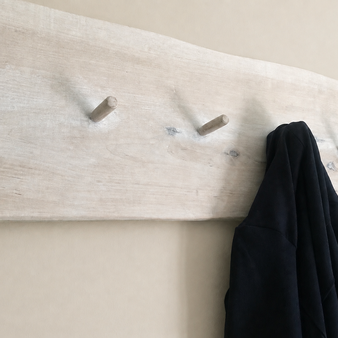 A rustic white-washed wooden coat rack with five pegs mounted on a beige wall, with a dark navy coat and a beige plaid scarf hanging from it.