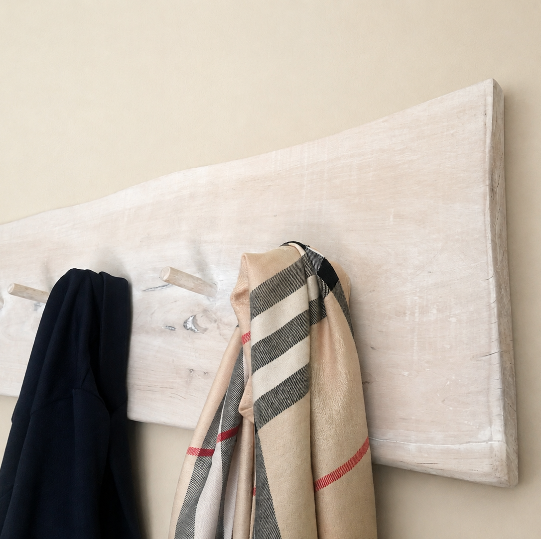 A long, rectangular light wood coat rack mounted on a beige wall, with several garments hanging from its hooks, including a dark navy jacket and a beige striped scarf.