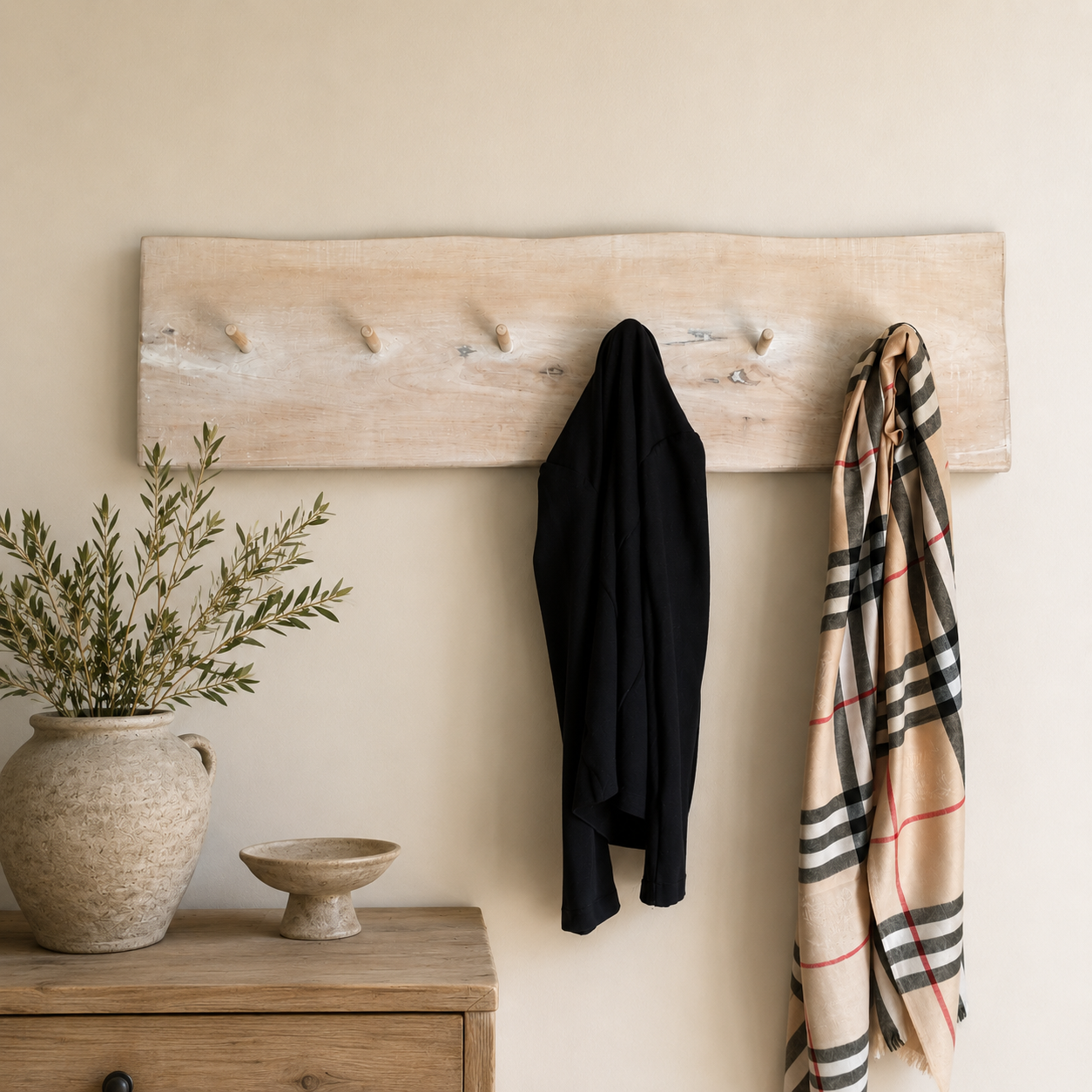 A rustic light wood wall-mounted coat rack with five pegs, shown hanging in a styled interior. A black coat is draped over the center peg and a beige plaid scarf hangs on the right.