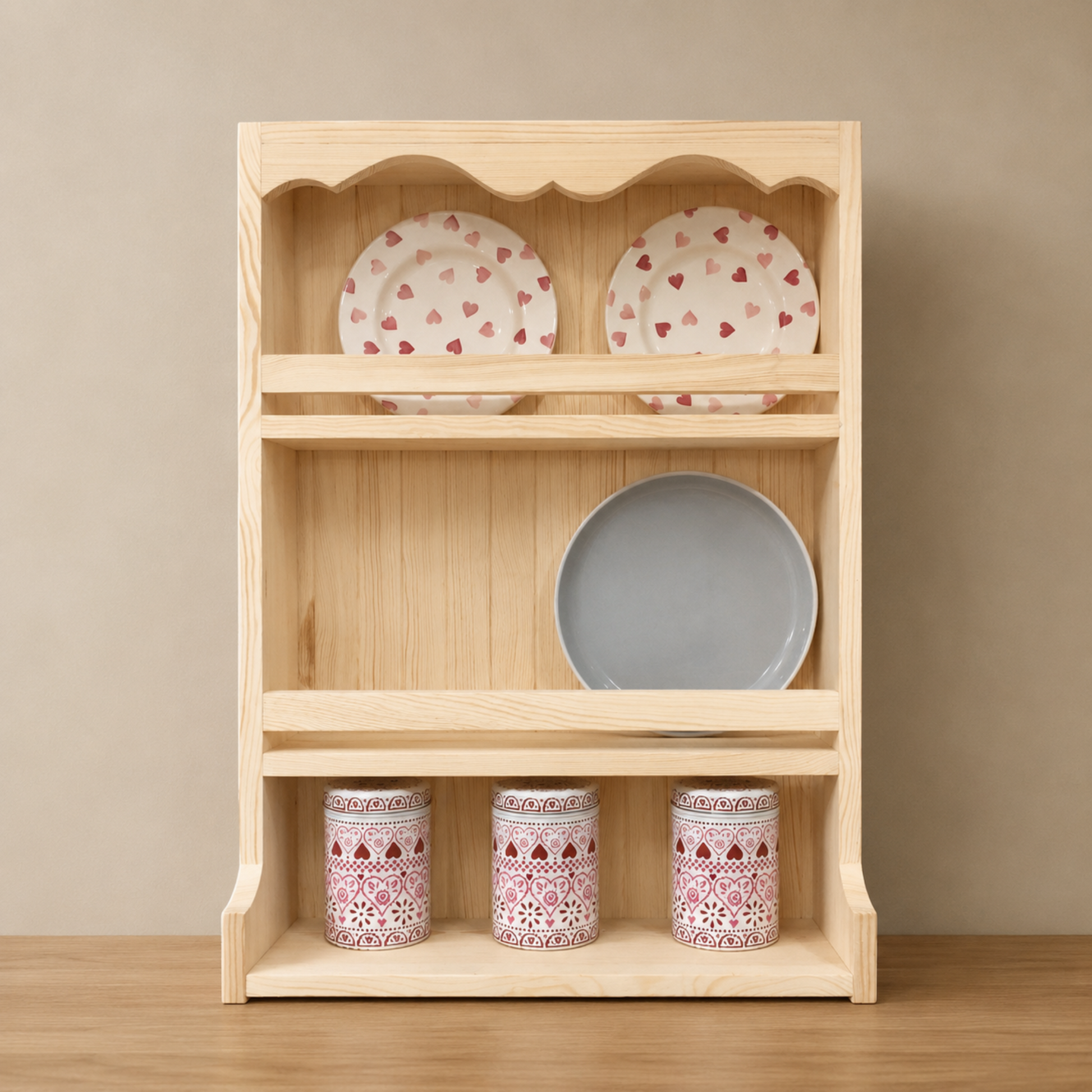 A light natural wood three-tier open shelf with scalloped top trim, shown holding two patterned plates on the top shelf, one gray plate on the middle shelf, and three matching patterned mugs on the bottom shelf, set against a neutral beige background.