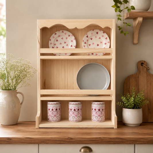 A light natural wood three-tier dish rack shelf styled on a wooden kitchen counter, holding two patterned plates, a larger plate, and three patterned canisters, with potted plants and a cutting board in the background.