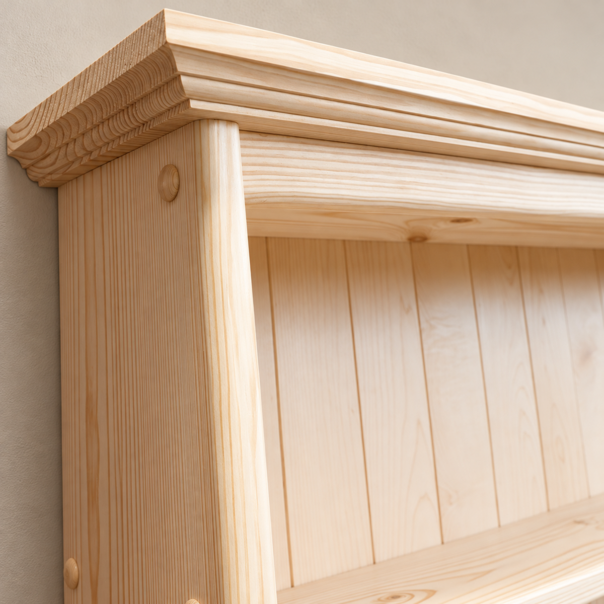 Close-up of a light natural wood wall shelf or cabinet corner with a decorative molded top, vertical slat back panel, and visible wood grain against a plain beige wall.