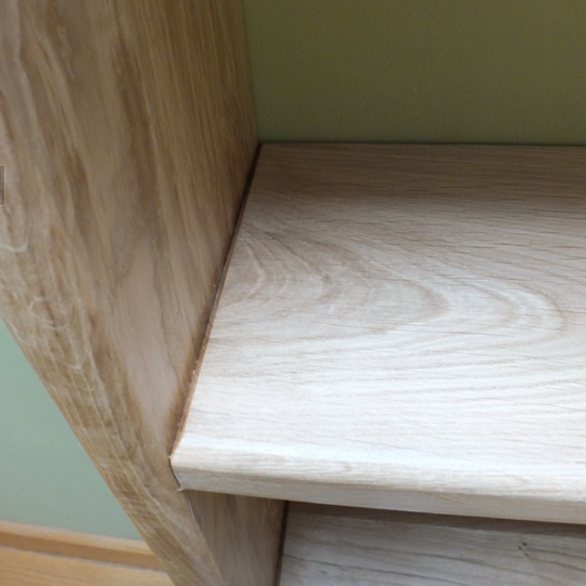 Close-up of a light natural wood shelf or cabinet corner with a pale green wall behind it.