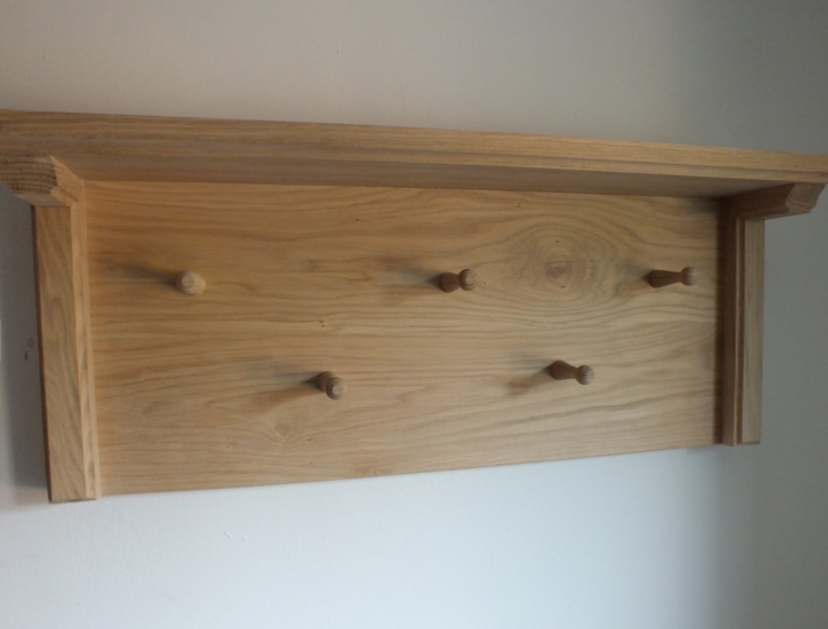 A light natural wood wall-mounted coat rack with a shelf and five wooden pegs, shown against a plain off-white wall.