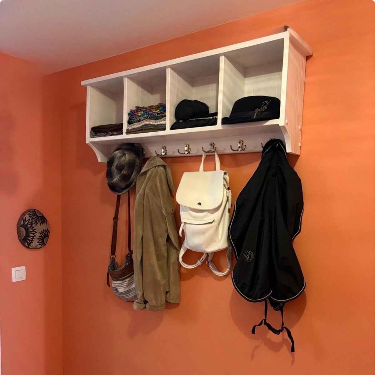 A white wall-mounted storage shelf with five cubbies and hanging hooks, shown in a real room with an orange wall. Hats, a bag, a jacket, and a backpack are hanging on the shelf and hooks.