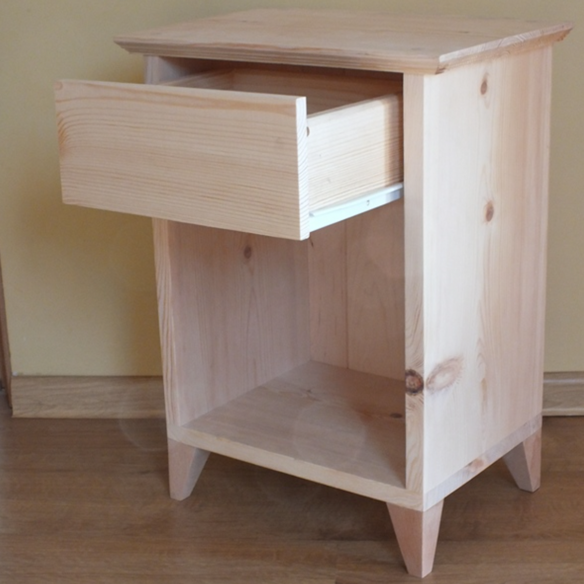 A light natural pine nightstand with a single pull-out drawer and an open lower shelf, shown on a wooden floor against a beige wall.