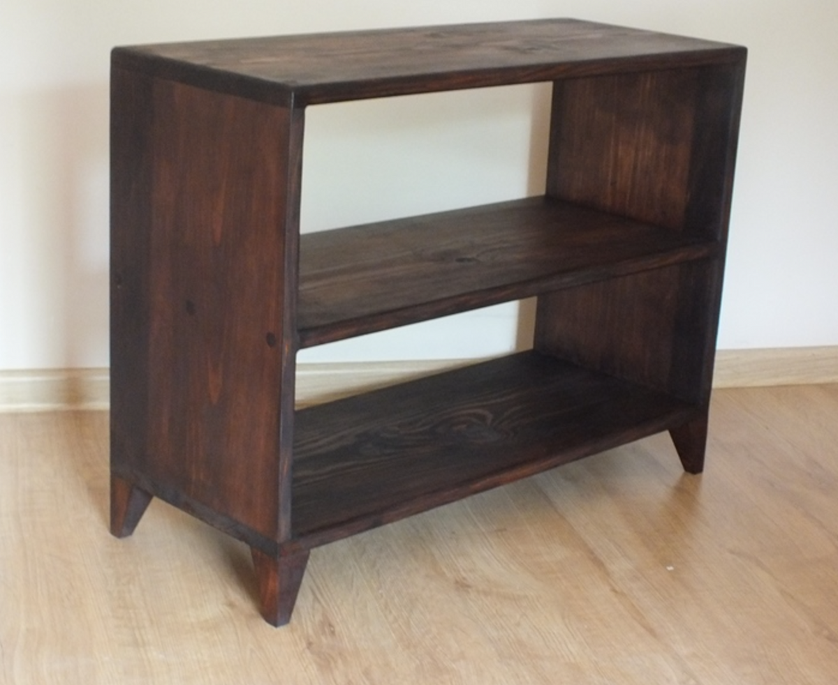 A dark brown wooden two-tier bookcase with tapered legs, shown standing on a light wood floor against a plain off-white wall.