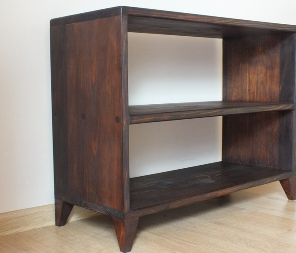 A dark brown wooden bookshelf with three open shelves and tapered legs, photographed in a simple indoor setting with a light wall and wood floor.