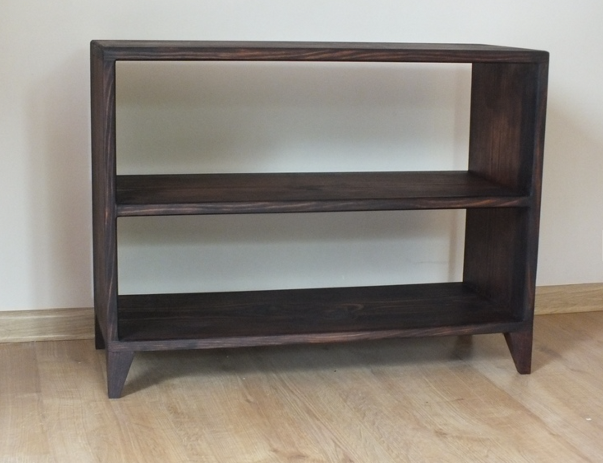 A dark brown wooden two-tier bookcase with open shelves and tapered legs, shown against a plain beige wall and light wood floor.