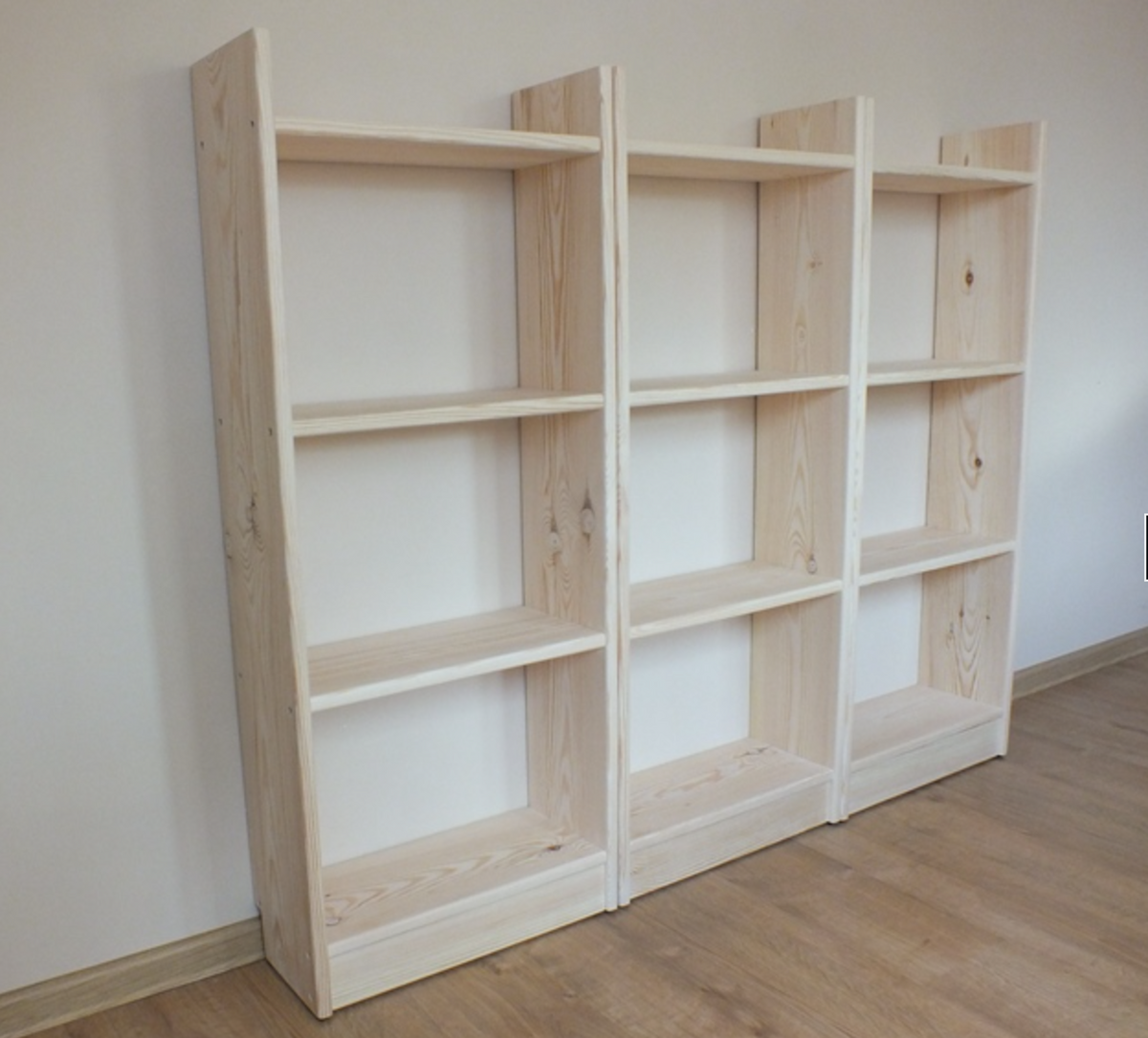 Three light natural wood bookshelves with five shelves each, arranged side-by-side against a plain off-white wall on a wooden floor.