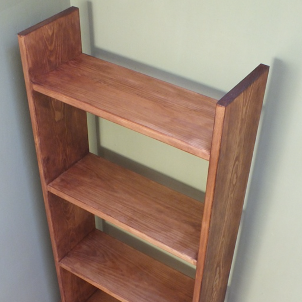 A four-tier wooden bookshelf with a warm medium-brown finish and visible wood grain, positioned in a room corner against a sage green wall and light wood floor.