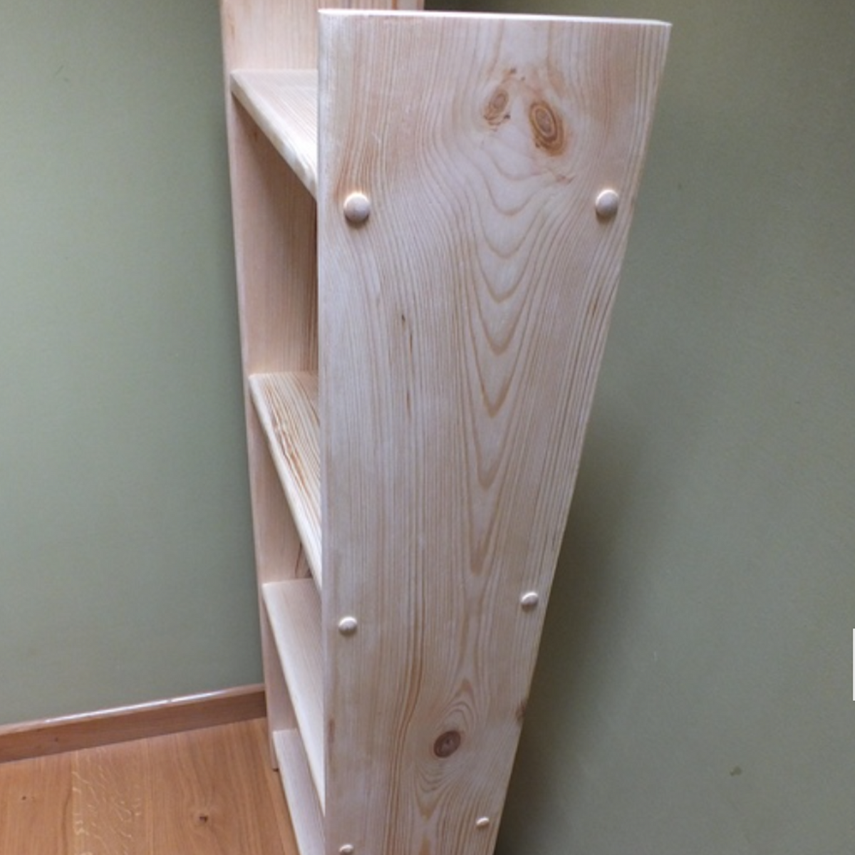 A light natural wood bookshelf with three open shelves and visible metal corner brackets, photographed standing in a room corner against a sage green wall and light wood floor.