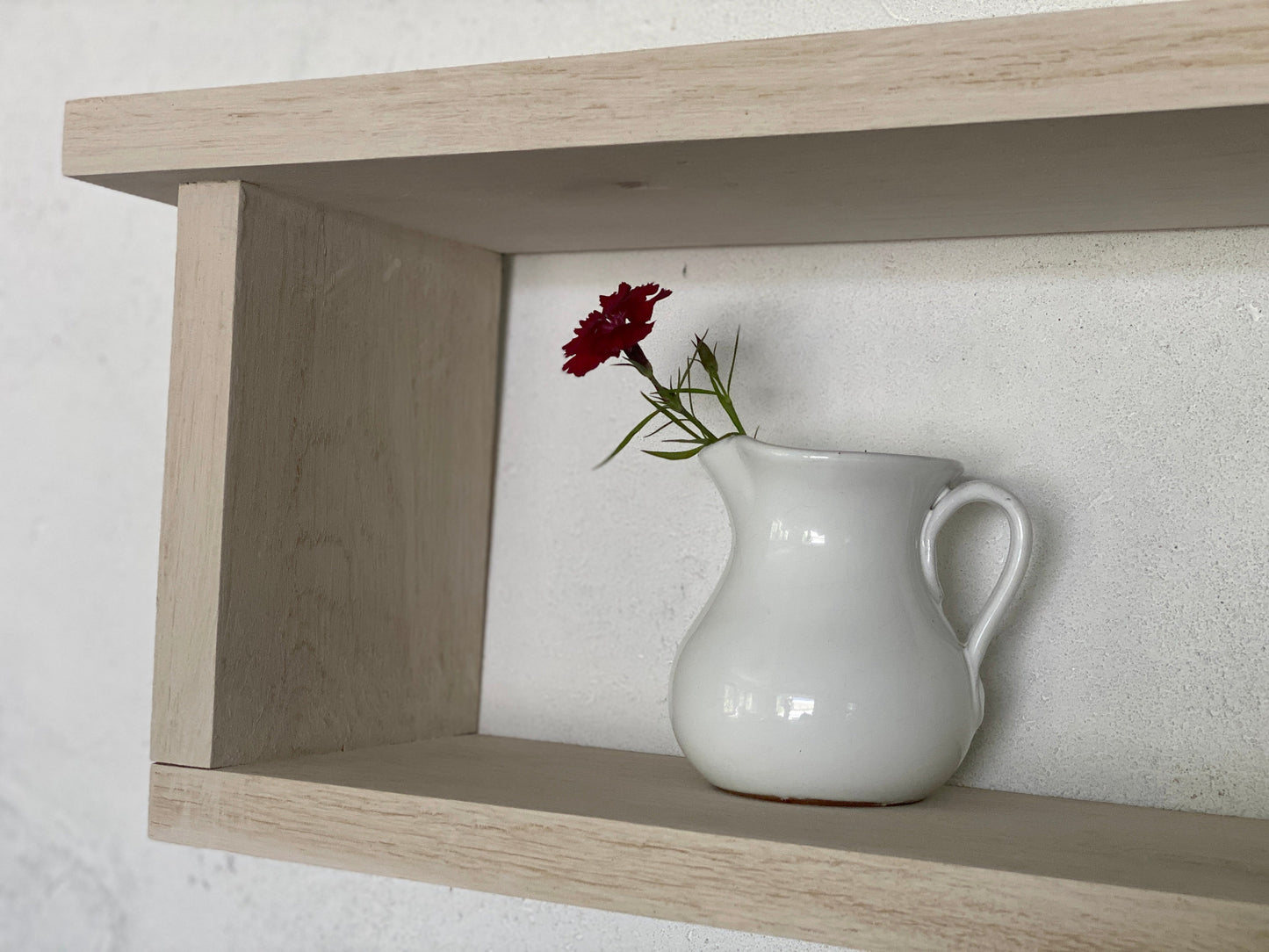 White ceramic pitcher with a red flower on a wooden shelf against a white wall.