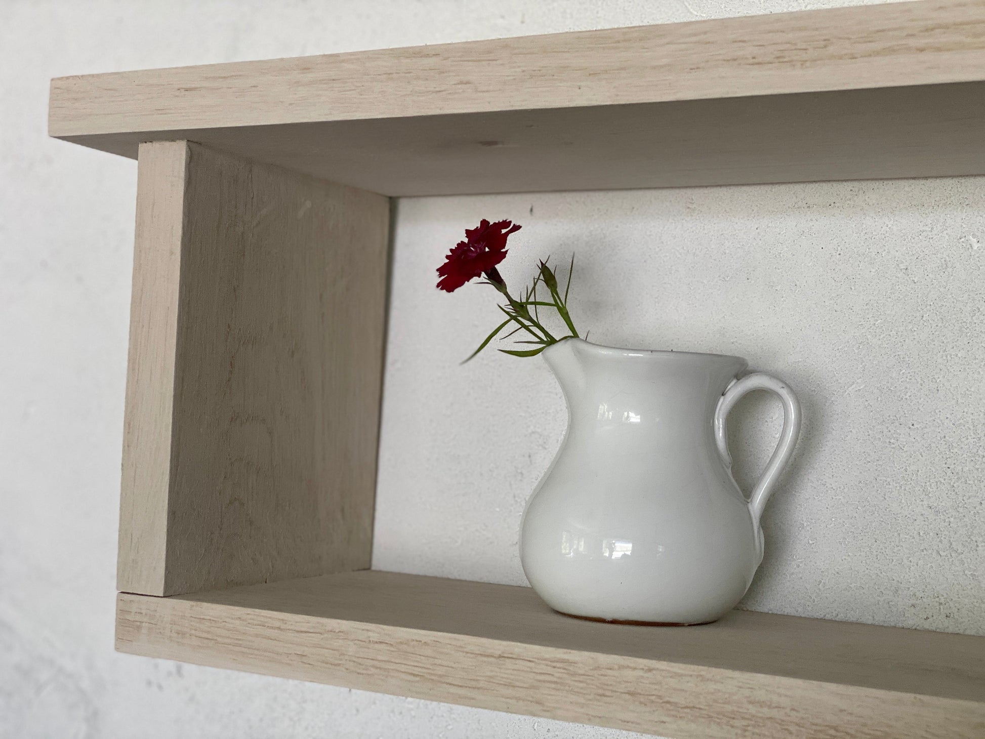 White ceramic pitcher with a red flower on a wooden shelf against a white wall.